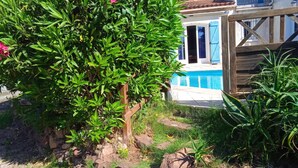 A poolside area with a wooden deck, a stone wall, and a house with blue shutters in the background. - Homerez - Charming Apt in Le Luc (Le Luc)
