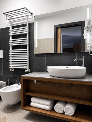 Un baño moderno con un lavamanos blanco tipo recipiente, un inodoro empotrado, un bidé, un perchero para toallas y un espejo con marco de madera.