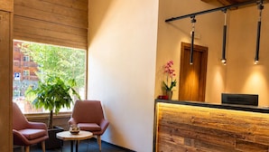 A reception area with a wooden reception desk, two chairs, a small table with a bowl of fruit, a potted plant, and a window with a view of buildings and trees. - Parc Hotel Posta (Marebbe)