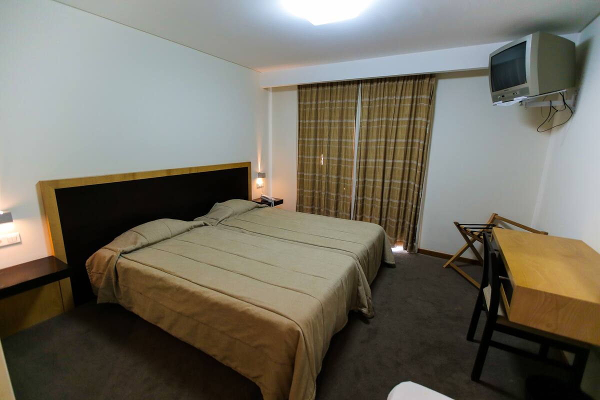 Una habitación de hotel con cama, escritorio, televisor y ventana con cortinas.