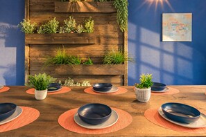 Un coin repas avec un mur en bois orné de plantes, un mur bleu avec un tableau, et une table en bois dressée avec des bols bleus et des sets de table orange.