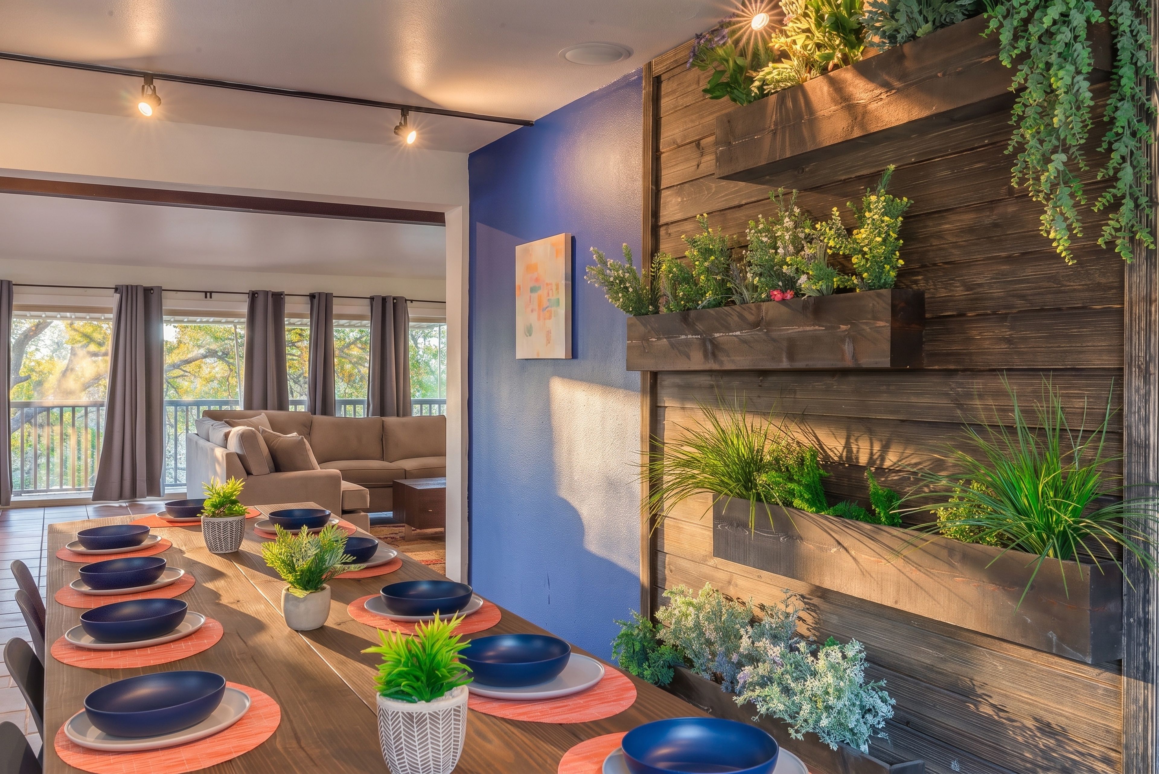 A modern dining area with a long table set for six, surrounded by a living room with a sofa and a view of the outdoors.