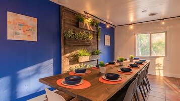 A dining area with a long wooden table set for a meal, surrounded by chairs, with a blue wall and a window providing natural light.