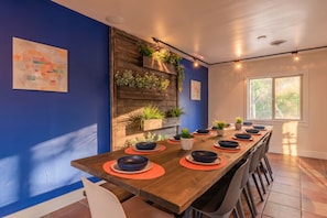 Uma sala de jantar com uma mesa de madeira comprida posta para refeição, rodeada por cadeiras, com parede azul e janela que oferece luz natural.