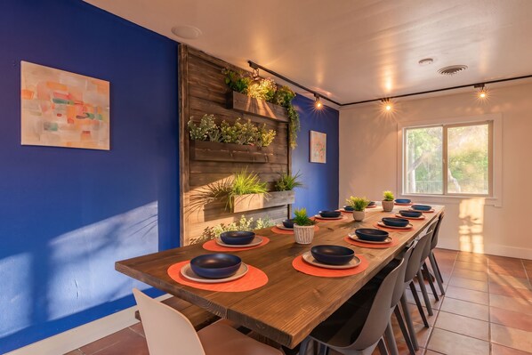 A dining area with a long wooden table set for a meal, surrounded by chairs, with a blue wall and a window providing natural light.