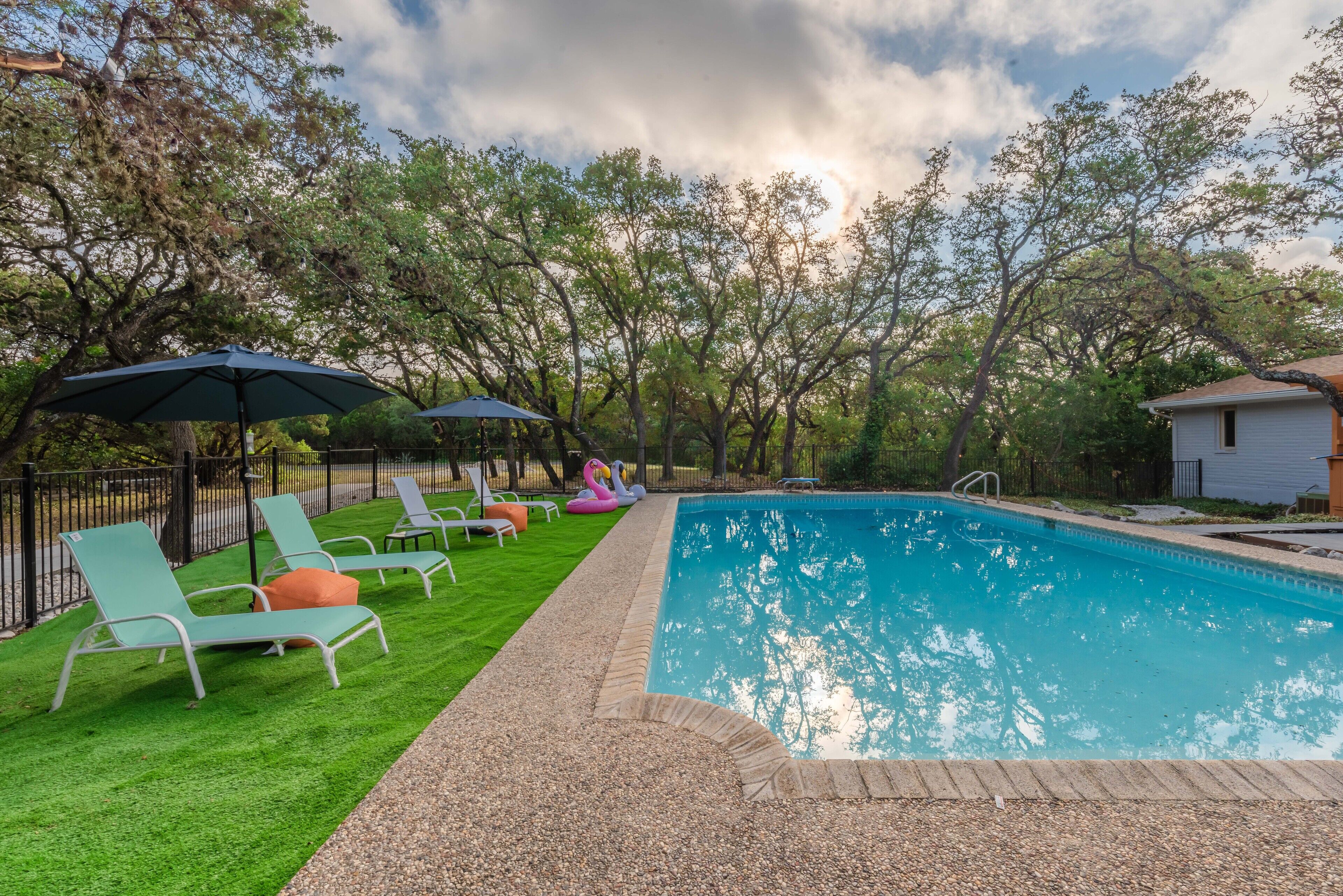 Uma piscina com espreguiçadeiras e guarda-sóis, cercada por árvores e uma pequena casa.