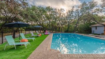 Uma piscina com espreguiçadeiras e guarda-sóis, cercada por árvores e uma pequena casa.