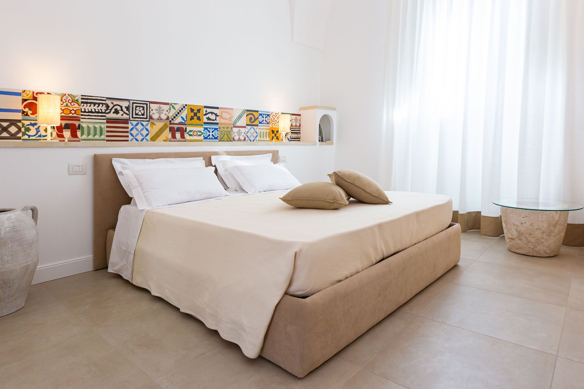 Un dormitorio ordenado con cama, almohadas, una mesita de noche y un estante en la pared con adornos.