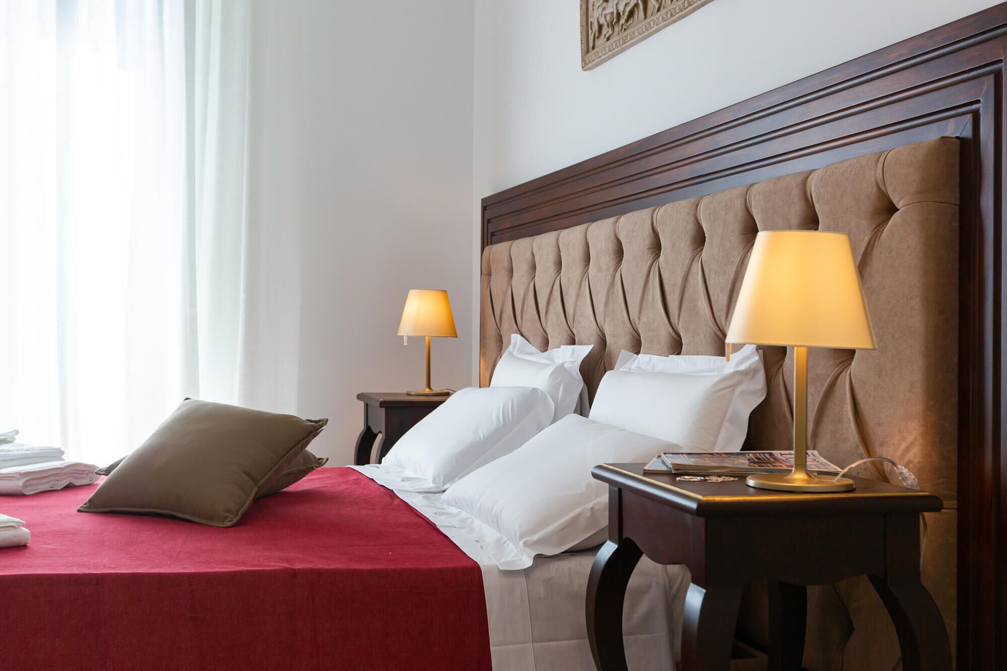 Habitación de hotel con una cama grande, dos mesitas de noche con lámparas, una colcha roja y un cabecero de madera.