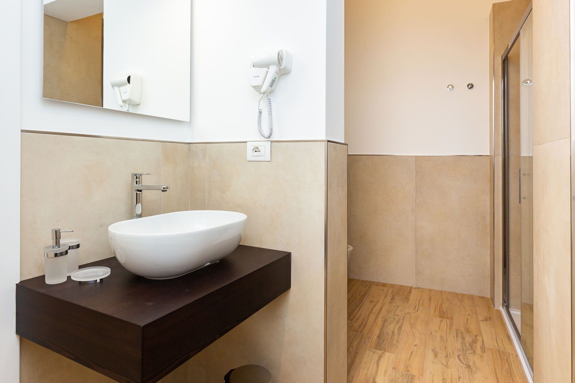 A modern bathroom with a large white sink, a dark wooden vanity, a mirror, a hairdryer, and a shower area.
