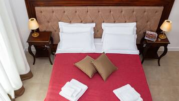 Una cama bien hecha con sábanas blancas, dos almohadas marrones y una colcha roja. Hay dos mesitas de noche con lámparas y una revista en una de ellas.