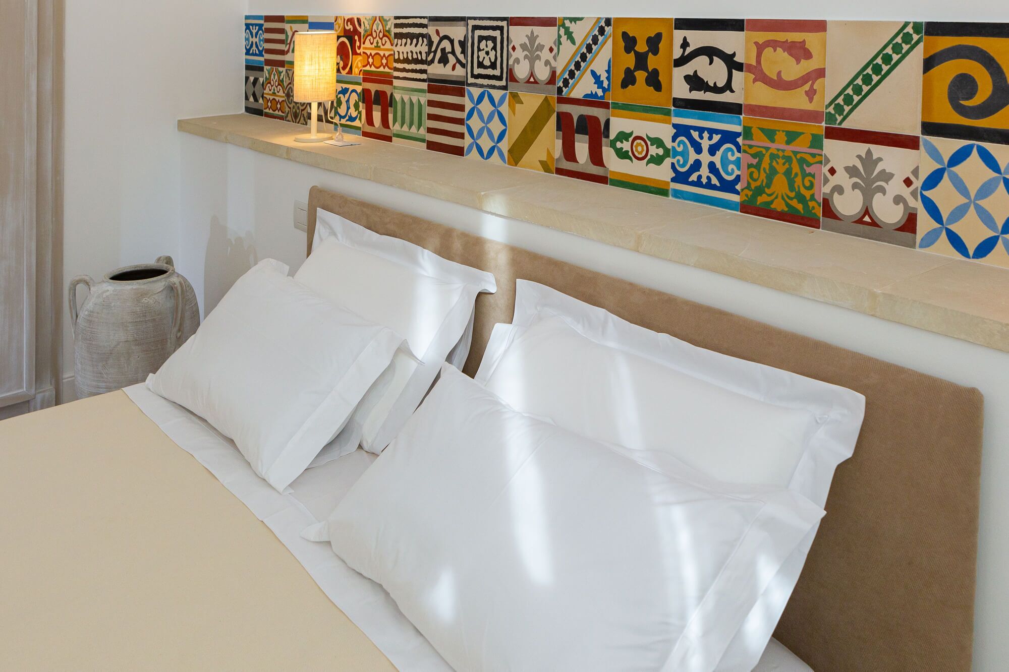 A neatly made bed with white linens and pillows, a headboard, and a wall with colorful patterned tiles.