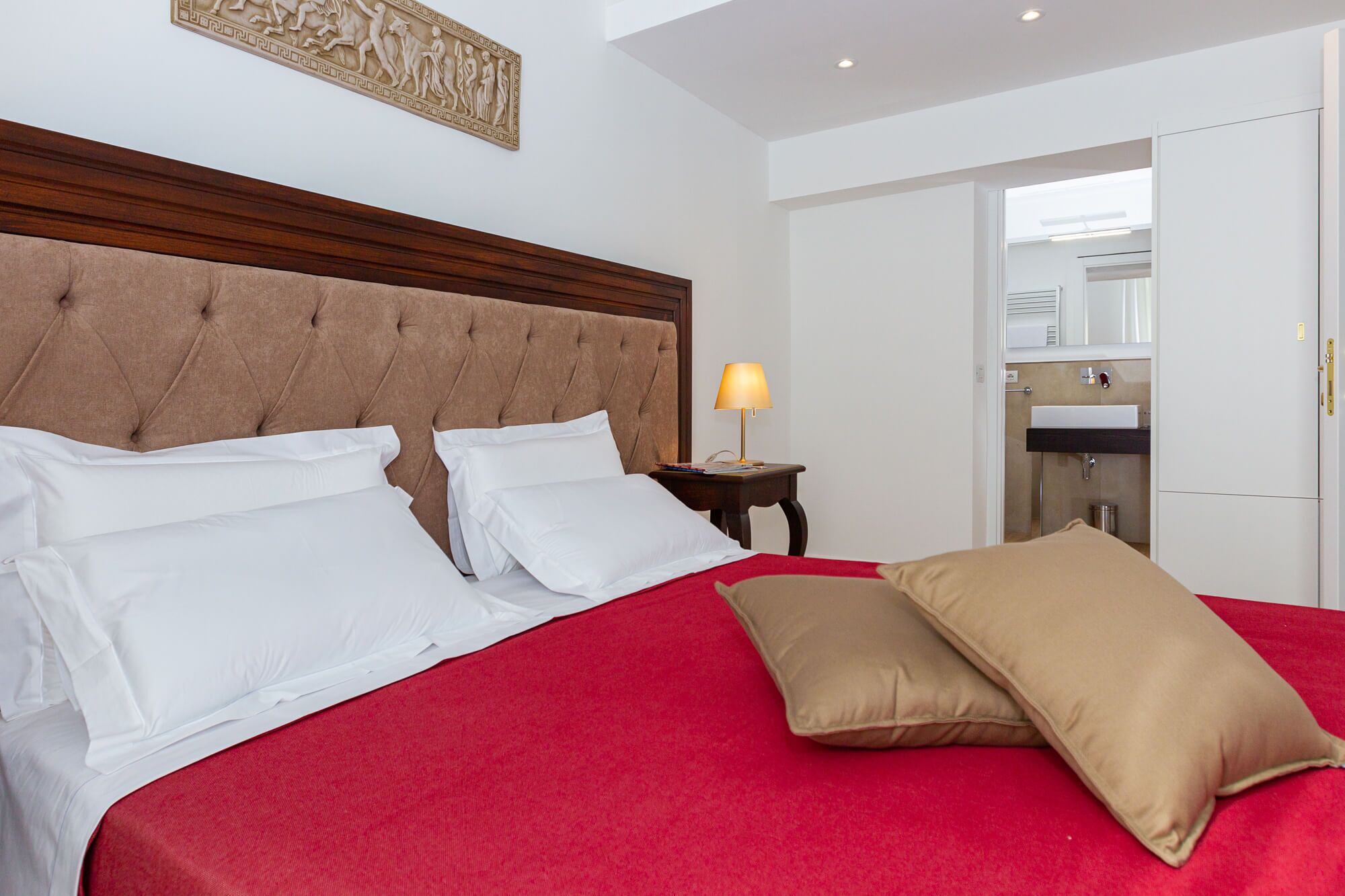 Una cama bien hecha con almohadas blancas y color beige, una colcha roja y un cabecero de madera. Hay una mesita de noche con una lámpara y un libro pequeño. Una puerta da acceso a un baño con espejo y lavamanos.