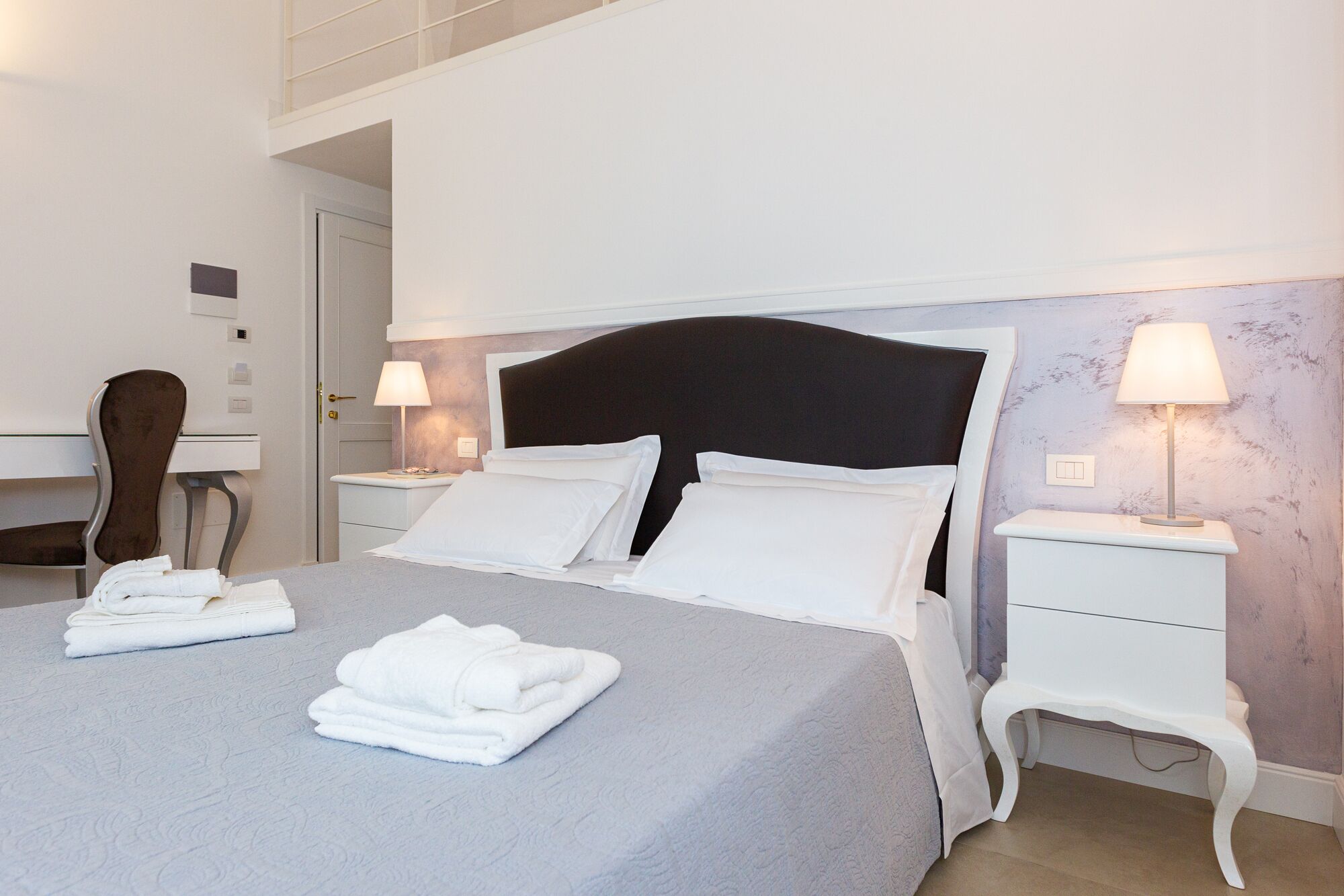 Un letto rifatto con lenzuola bianche, due comodini con lampade, una sedia e una scrivania in una moderna camera d'albergo.