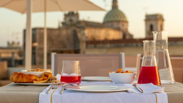 Une table dressée pour un repas, avec des croissants, un verre d’une boisson rouge et un saladier de fruits, donnant sur un bâtiment historique surmonté d’un dôme.