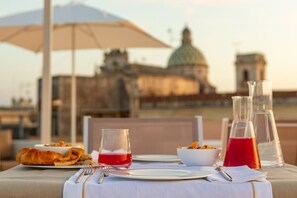 Mesa preparada para una comida con cruasanes, un vaso de bebida roja y un bol de fruta, con vista a un edificio histórico con cúpula.