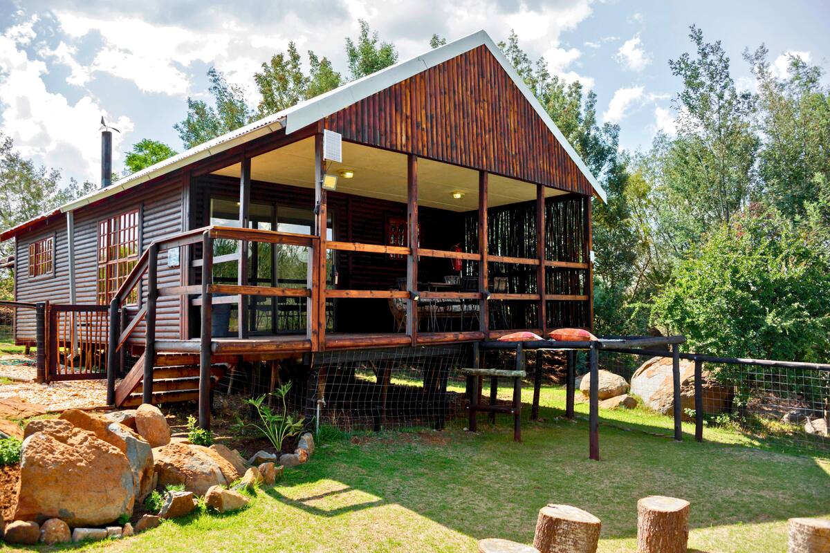 A rustic wooden cabin with a large porch, surrounded by rocks and trees.