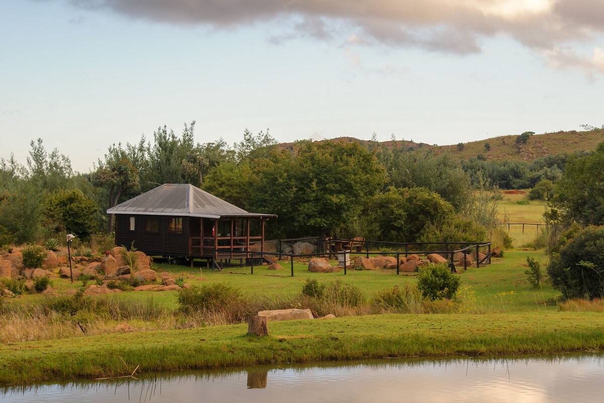 A rustic cabin with a metal roof, surrounded by a wooden deck and a fenced area with rocks.