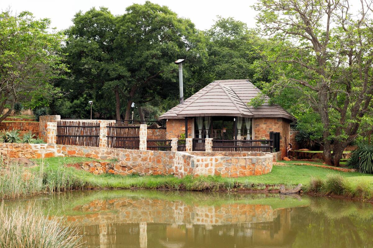 A stone building with a tiled roof, surrounded by a stone wall and a fence, near a pond with reflections.