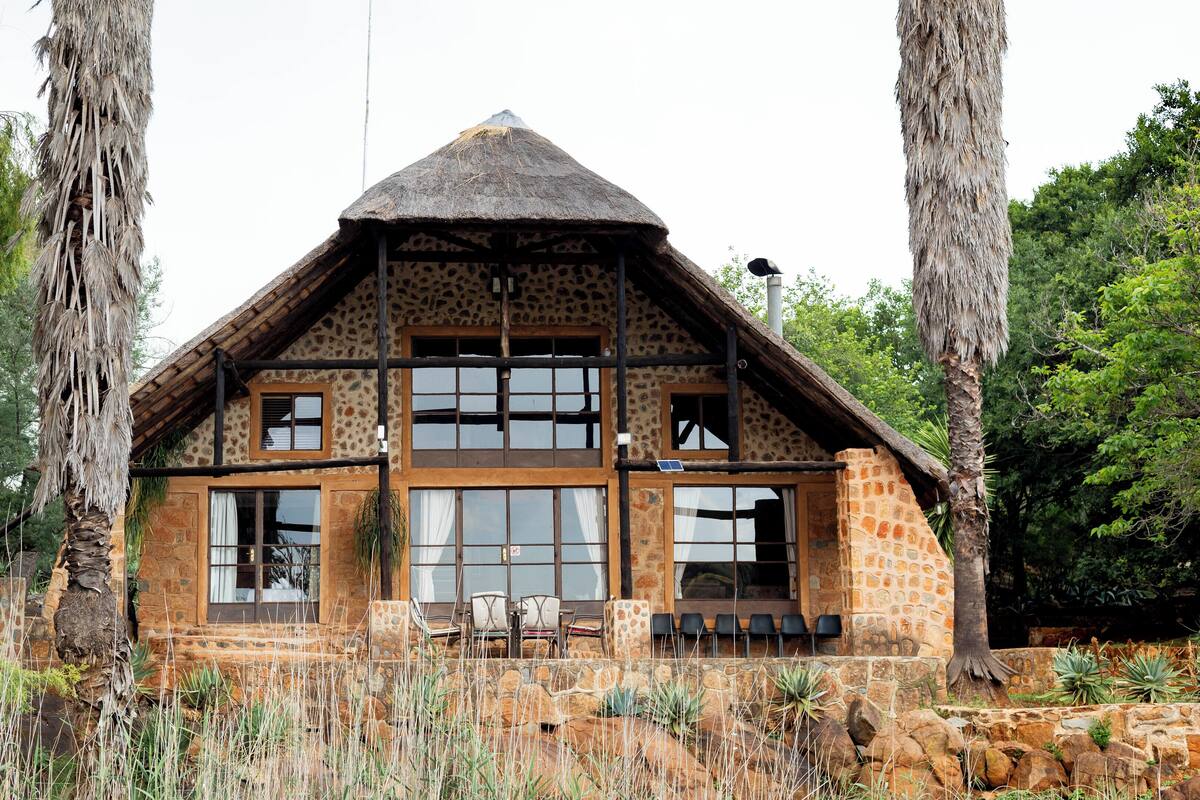 A stone house with a thatched roof, large windows, and a balcony with chairs.