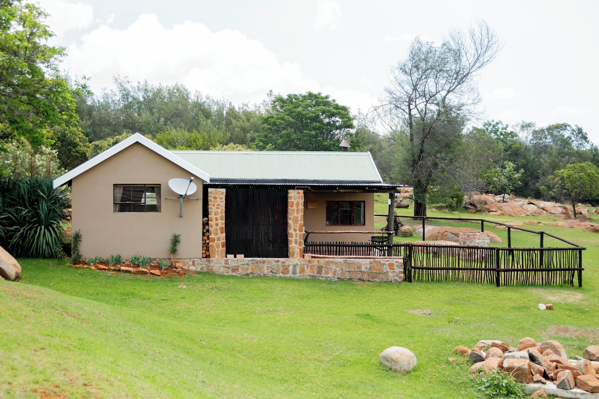 A single-story house with a stone foundation and a green metal roof, surrounded by a grassy yard and a stone wall.