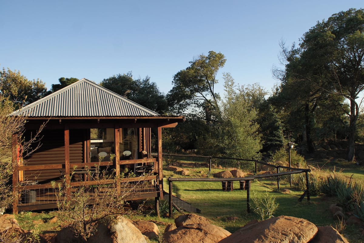 A rustic wooden cabin with a corrugated metal roof, surrounded by a natural landscape with trees and rocks.