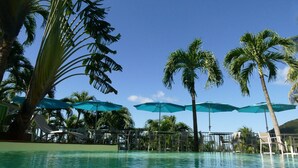A pool area with clear blue water, surrounded by palm trees and blue umbrellas. - "O Coeur de Deshaies" (Deshaies)