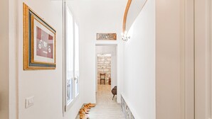 A hallway with a framed picture, a sign, and a row of shoes leading to a room with a stone wall and a chair. - Palazzo dei Normanni Stylish Apartment (Palermo)