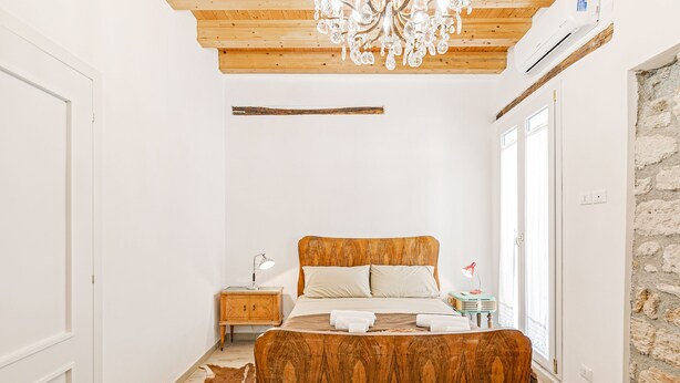 A bedroom with a wooden bed, a chandelier, a stone wall, and a large window. - Palazzo dei Normanni Stylish Apartment (Palermo)