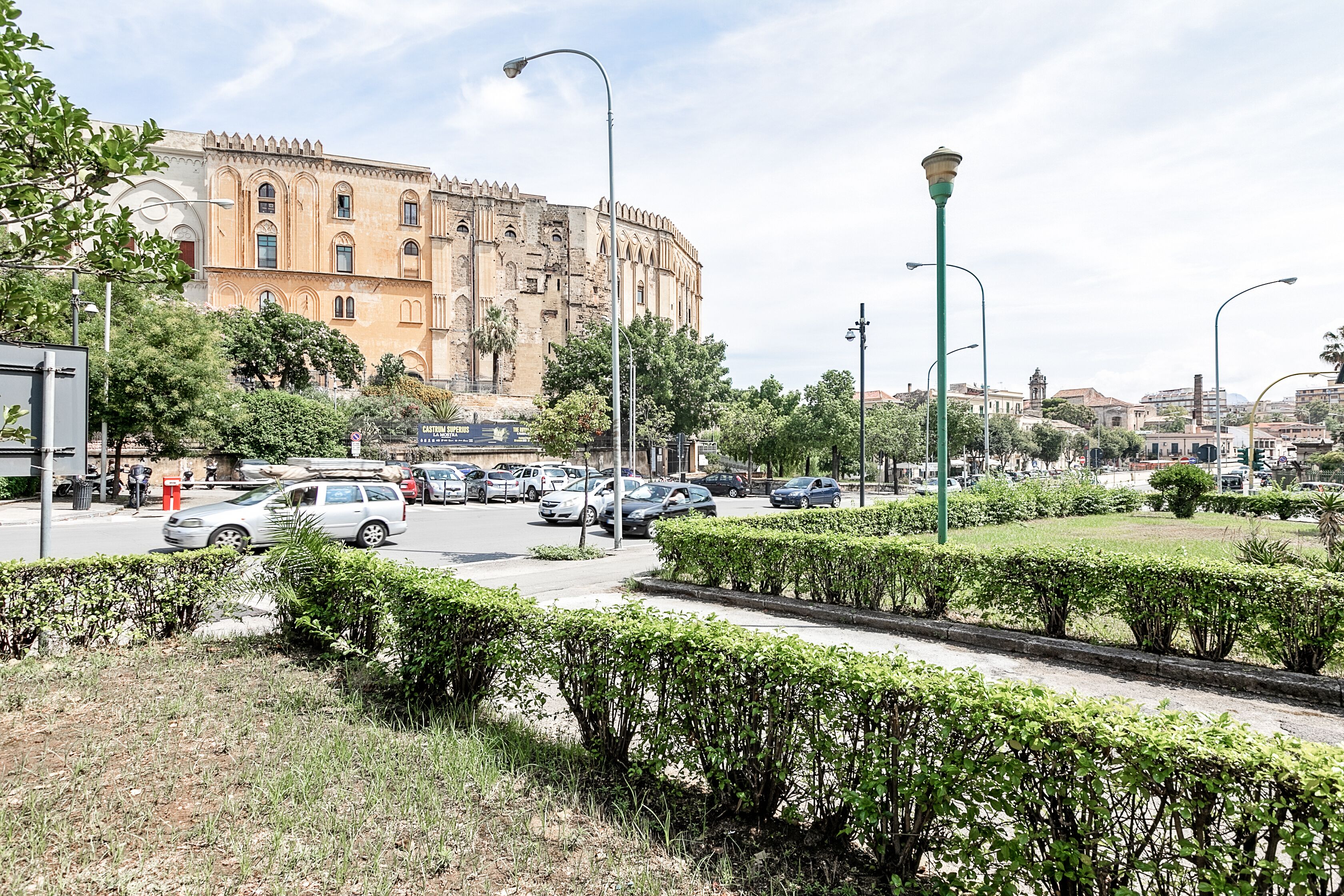 Une rue bordée de voitures garées, un grand bâtiment doté de fenêtres en arc de cercle et un beau buisson taillé au premier plan.