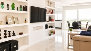 Ein modernes Wohnzimmer mit einem Flachbildfernseher, weißen Regalen mit Dekoration, einem beigen Sofa und Blick durch große Fenster.