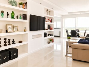 A modern living room with a flat-screen TV, white shelving with decorative items, a beige sofa, and a view through large windows.