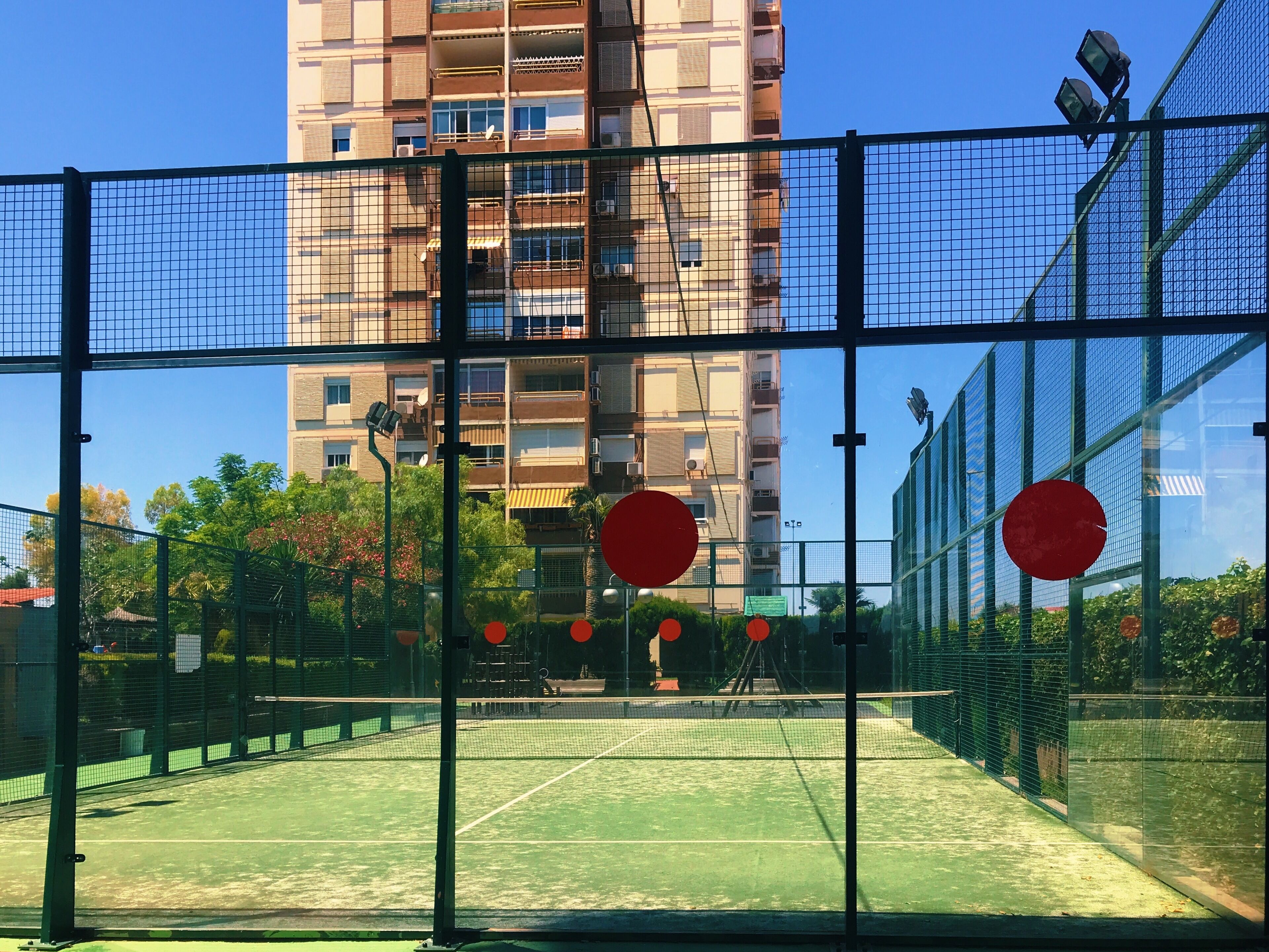 Una cancha de tenis con red, rodeada por una valla verde, y un edificio alto al fondo.