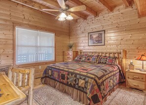 A wooden cabin bedroom with a large bed, a wooden headboard, a nightstand with a lamp, a window with blinds, and a ceiling fan. - Luxury! 2 Master Suites w/Fireplaces|Game Room (SEVIERVILLE)