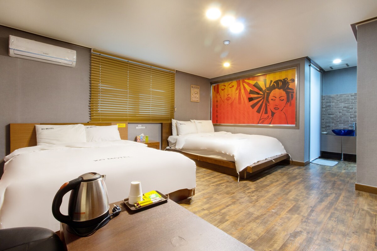 Habitación de hotel con dos camas, cafetera, bandeja con tazas y una botella, un cartel de 'Se busca' y una obra de arte grande en la pared.