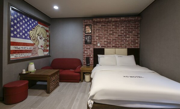 Habitación de hotel con cama, una silla roja, una mesita, un cubo de basura y una pared con diseño de ladrillo y un cuadro enmarcado.