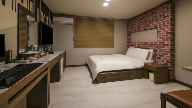 Un dormitorio con escritorio, cama, pared decorada con ladrillos y ventana con persianas.