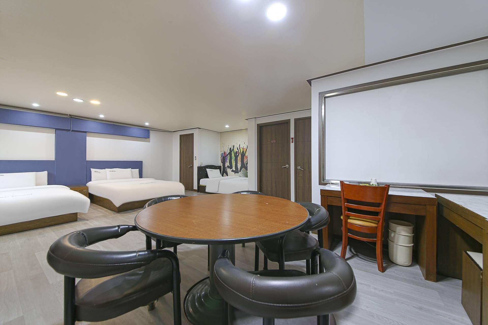 A hotel room with a large round table, two beds, a desk with a chair, and a whiteboard.