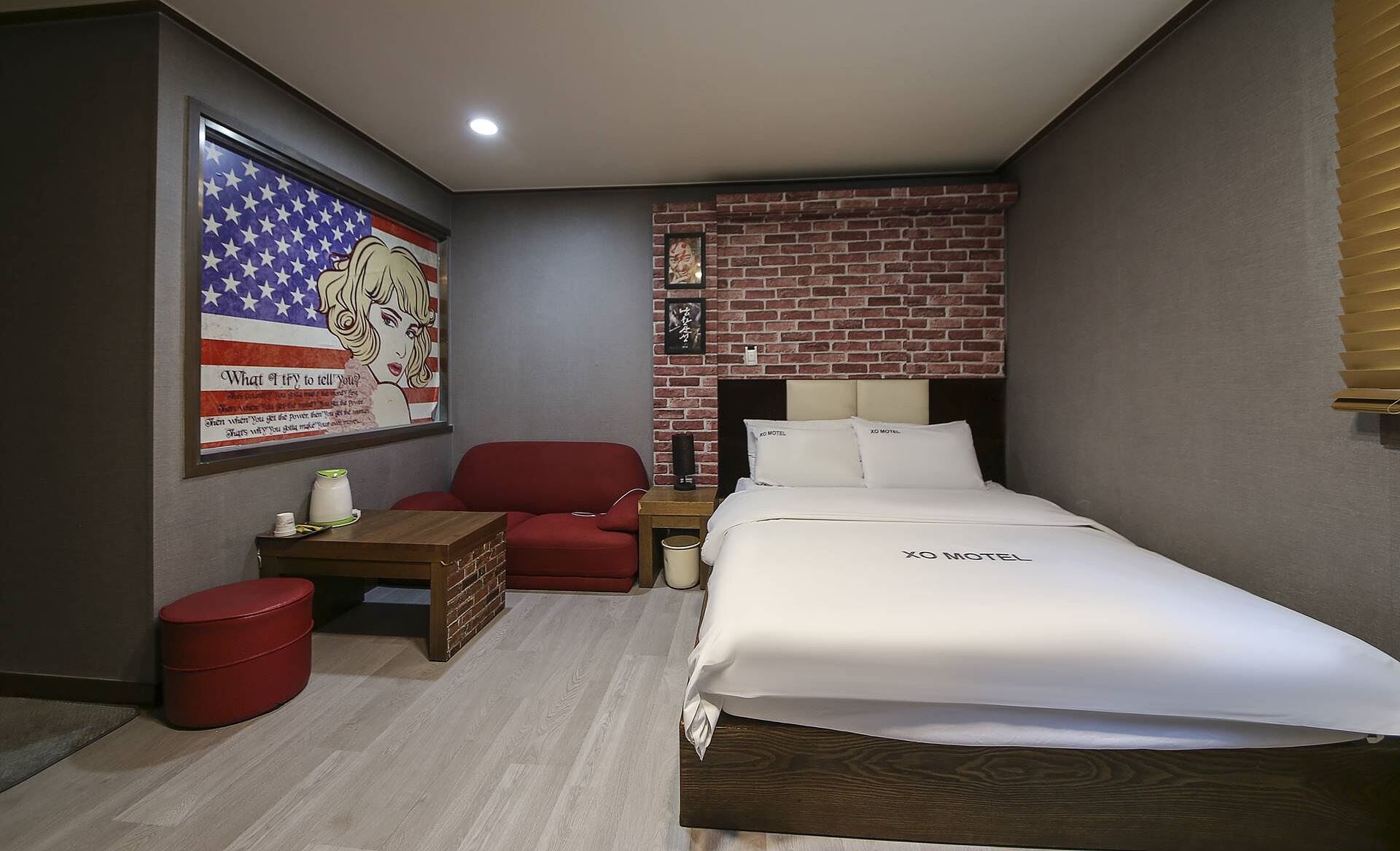 A hotel room with a bed, a red chair, a small table, and a wall with a brick pattern.