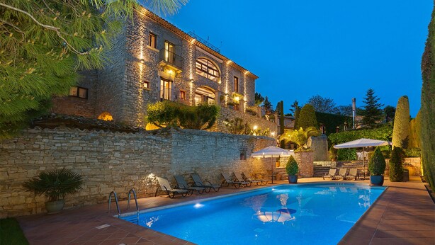 A stone building with a swimming pool, outdoor seating, and a well-lit balcony. - Cal Carulla (Pujalt)