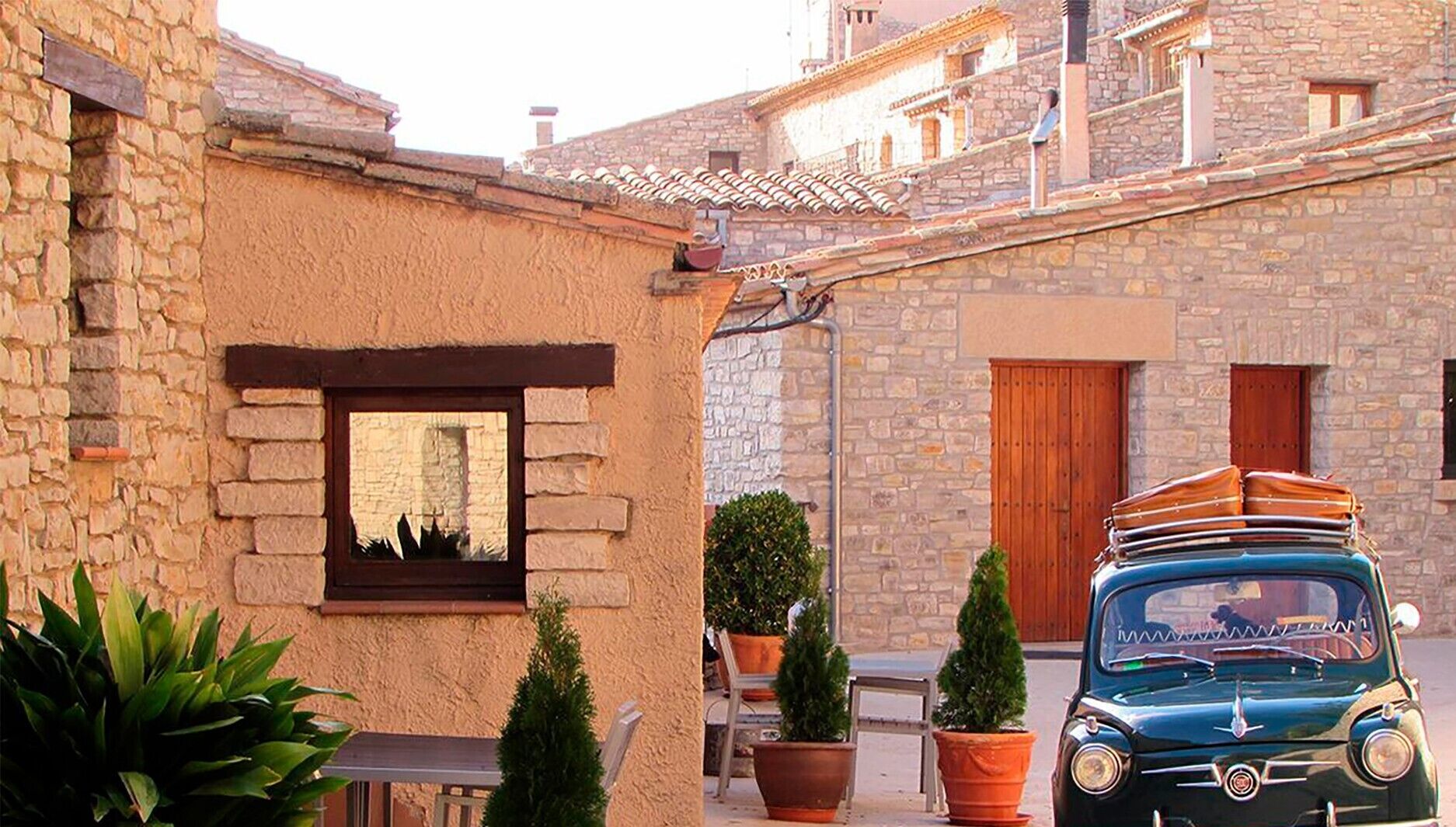 Un edificio de piedra con una ventana y una puerta, un coche clásico estacionado y plantas en macetas.