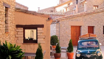 Un edificio de piedra con una ventana y una puerta, un coche clásico estacionado y plantas en macetas.
