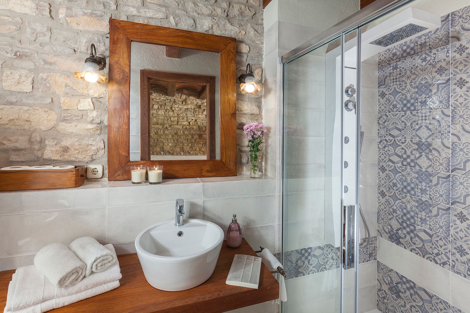 Un baño con pared de piedra, tocador de madera, lavamanos blanco, cabina de ducha de vidrio y azulejos estampados.