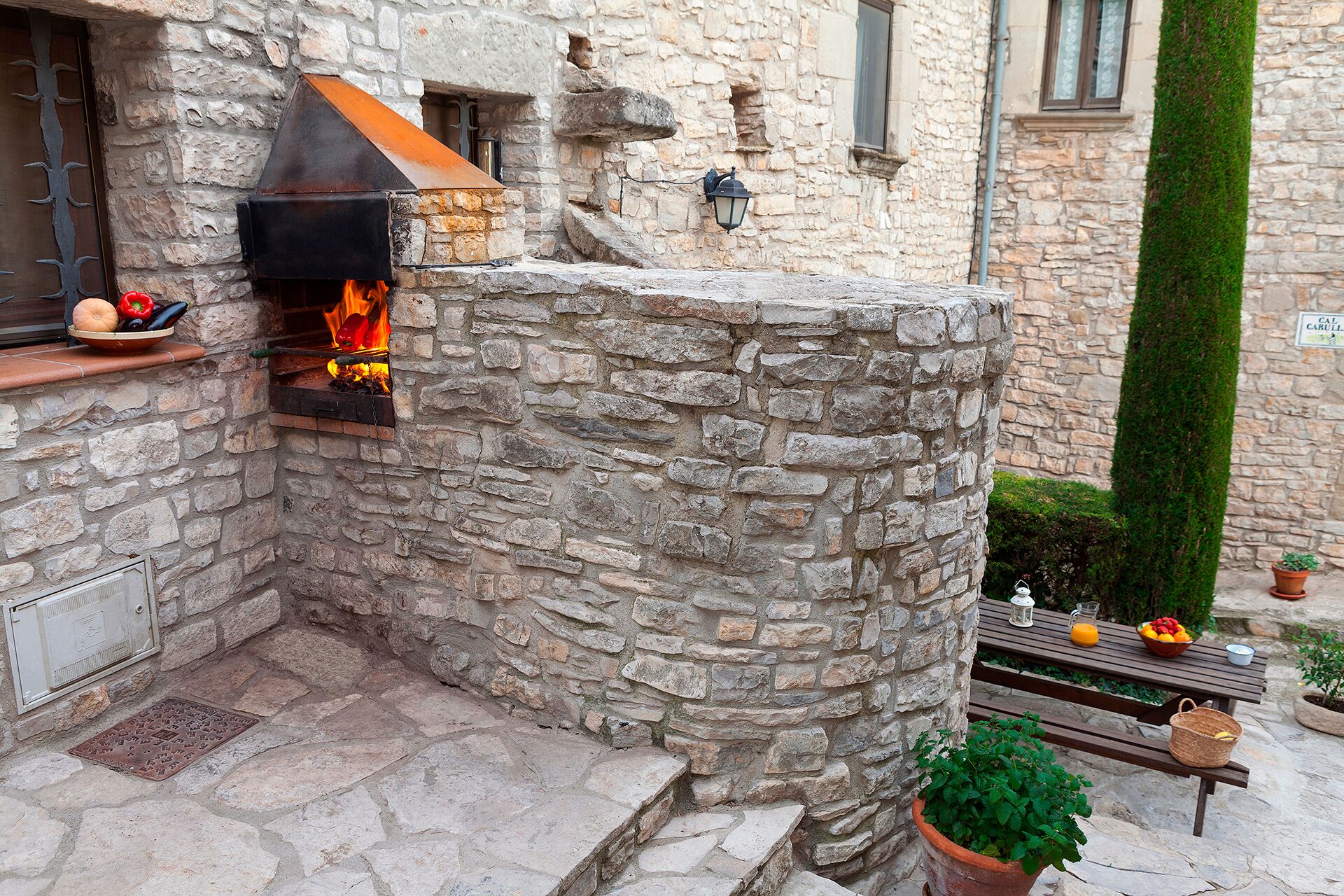 Une cuisine extérieure en pierre, dotée d’un poêle à bois, d’un mur en pierre et d’un banc avec une table.