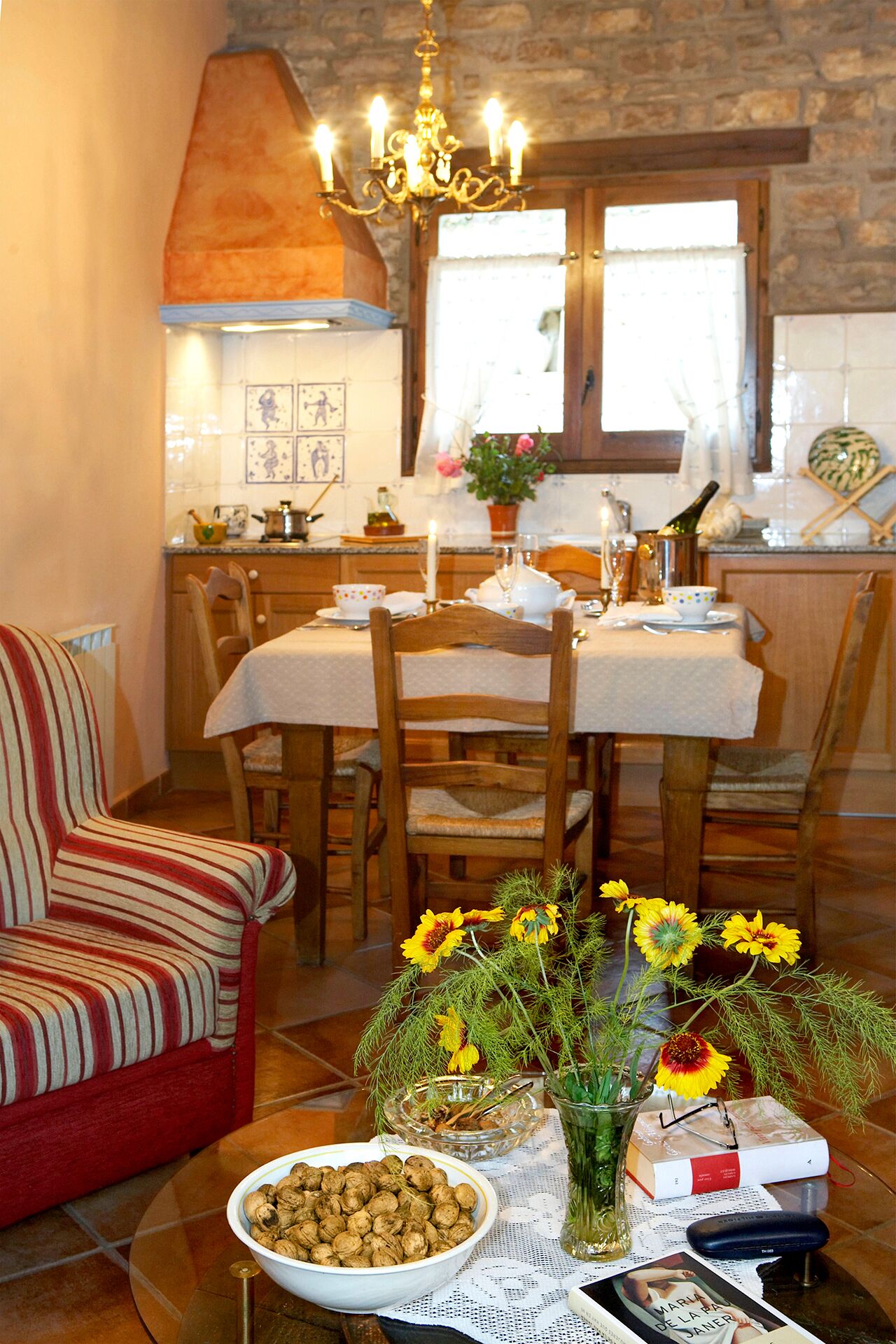 Un comedor con una mesa de madera dispuesta para cuatro personas, un sillón a rayas y un jarrón con flores amarillas.