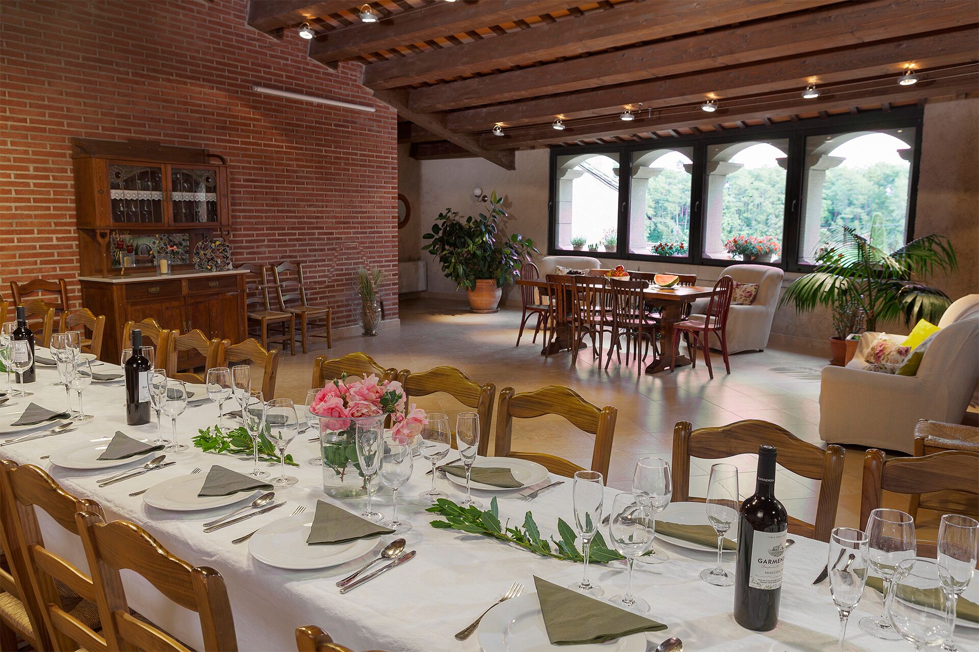 A dining area with a long table set for a meal, surrounded by wooden chairs. There are potted plants and a view of greenery through large windows.