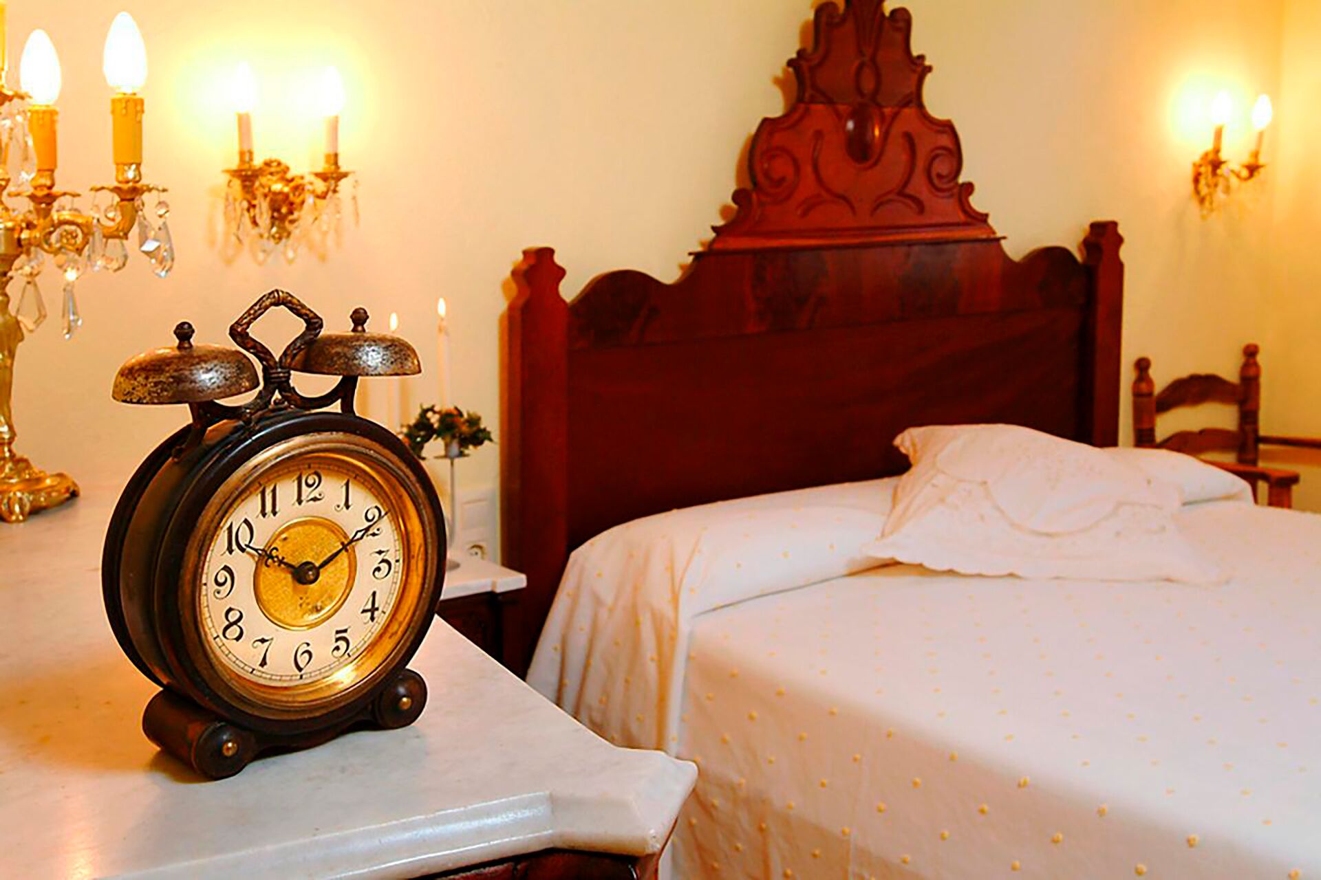 Een klassiek houten bed met witte beddengoed, een vintage wekker op een nachtkastje, en wandklemmen met kaarsen.