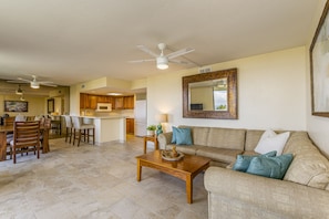 A spacious living area with a sofa, coffee table, and dining table. There is a kitchen in the background with wooden cabinets and a refrigerator. - Summer Deal White Sands Beach Condo with Pool Lanai and Ocean Views (Kailua Kona)
