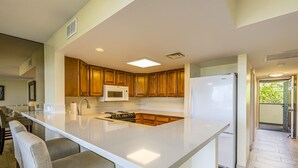 A modern kitchen with a large island, wooden cabinets, stainless steel appliances, and a view to the outside. - Summer Deal White Sands Beach Condo with Pool Lanai and Ocean Views (Kailua Kona)