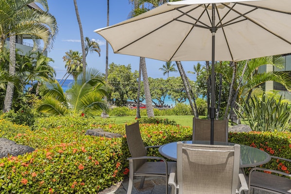 A tropical outdoor seating area with a table and chairs under a large umbrella, surrounded by lush greenery and palm trees. - Summer Deal White Sands Beach Condo with Pool Lanai and Ocean Views (Kailua Kona)